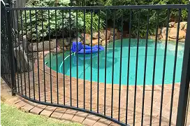 A black metal safety fence with vertical bars curving around a backyard swimming pool.