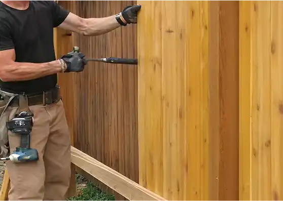 A worker wearing a tool belt uses a hammer to install a new vertical plank on a wooden privacy fence.