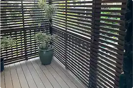 A modern dark brown privacy screen with horizontal slats on a deck, with a potted plant in the corner.