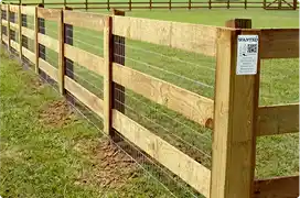 A wooden three-rail fence with wire mesh infill, installed in a green pasture.