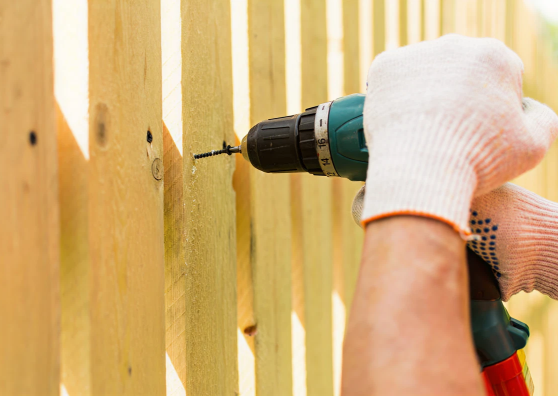 A worker wearing a tool belt uses a hammer to install a new vertical plank on a wooden privacy fence.
